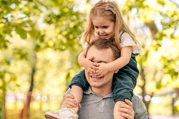  dad with little daughter play in the park on the street, toddler sits on his fathers shoulders, close-up, child laughs, closed eyes to dad, concept happy fathers day.