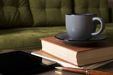 Tablet pc, stack of book and notebook, cup of coffee, pen on the wooden desk against green sofa.Empty space