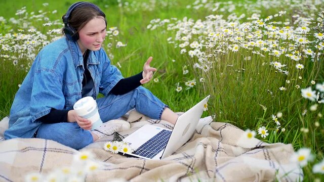 Teenage student girl sitting on lawn in park talking on video call with black headphones and waving one hand in other holding cup of coffee. Female wearing casual jeans and blue jaket working on air.