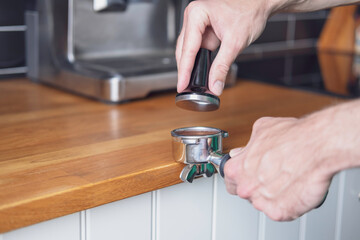 A man in the process of pressing coffee with a tamper on filter holder to make coffee in a carob coffee machine (coffee maker) in the kitchen at home near a wooden tabletop against a black wall.