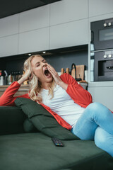 sleepy woman covering mouth with hand while yawning on sofa near tv remote controller