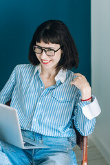 Beautiful brunette with a short haircut works in the home office on a blue background. Typing on a laptop, calling on the phone, transferring documents, making notes, drinking coffee from a mug.