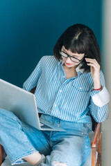 Beautiful brunette with a short haircut works in the home office on a blue background. Typing on a laptop, calling on the phone, transferring documents, making notes, drinking coffee from a mug.