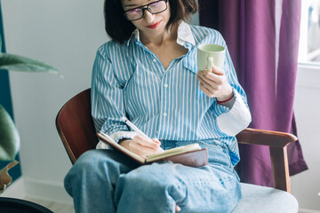 Beautiful brunette with a short haircut works in the home office on a blue background. Typing on a laptop, calling on the phone, transferring documents, making notes, drinking coffee from a mug.