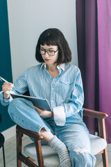 Beautiful brunette with a short haircut works in the home office on a blue background. Typing on a laptop, calling on the phone, transferring documents, making notes, drinking coffee from a mug.