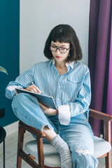 Beautiful brunette with a short haircut works in the home office on a blue background. Typing on a laptop, calling on the phone, transferring documents, making notes, drinking coffee from a mug.