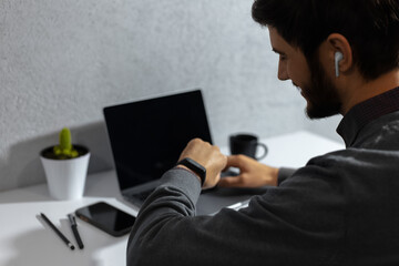 Close-up portrait of young smiling businessman verifies time on his smartwatch, using wireless earphones. Sitting in office beside workplace.