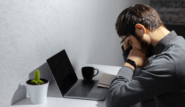 Portrait of sad businessman setting beside work place with laptop, coffee mug and cactus on desk. Using wireless earphones and smartwatch.