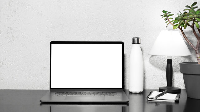 Freelance Business Workplace. Laptop With Mockup On Black Desk Beside Steel Thermo Water Bottle, Lamp, Flower And Notebook With Pen. Textured Wall Background.