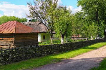 Fototapeta na wymiar Typical rural street in ancient Ukrainian village in sunny spring day. Clay house under thatched roof with a garden surrounded by a wicker fence. Concept of historical buildings of ancient Ukraine