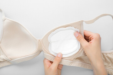 Young mother hand putting white breast pad in bra on light gray table background. Clothes...