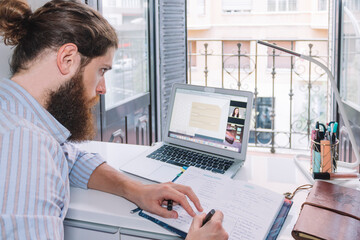 Bearded man on a video call writing in a notebook in front of the window