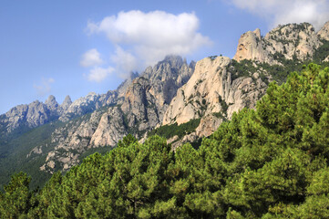 Pins Laricio et Aiguilles de bavella en Corse, sur fond de ciel bleu nuageux et nature sauvage et abrupte des rochers formant une montagne découpée au profil accidenté et dentelé.