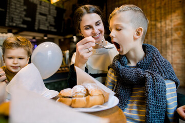 Beautiful young woman with her baby sons in cafe, having breakfast with fresh croissants, desserts and coffee, sitting at the wooden table. Happy family eating together, mom feeds her son