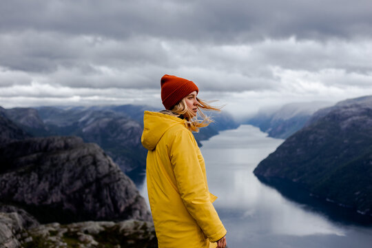 Dreamy blonde woman with wild hair in yellow raincoat and mustard hat in profile on background of Lysefjord with grey mountains and low cloudy sky in Norway