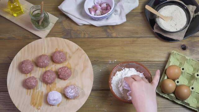 a female cook cooks meatballs, dipping balls of minced meat in flour on a wooden table with all the ingredients.