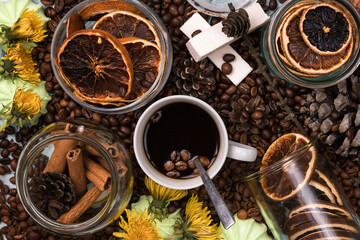 White Cup of coffee on coffee beans. Yellow dandelions. Light wooden table. Fir and cedar cones on the table. Green marshmallows and white marshmallows. Dried citrus. Brown background.