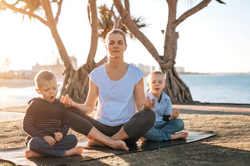 Woman meditating outdoors in park with two cute little sons. The ocean on background. Selective focus.