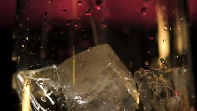 Pouring fresh alcoholic drink into the glass with ice cubes on dark background with red dim light. Stock footage. Close up of process of cocktail preparation, adding beverage into the glass.