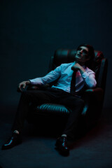 Portrait of a man in blue and red contrasting lighting in a white shirt with a tie and a facial injury, posing sitting in a leather chair. Futuristic neon lighting