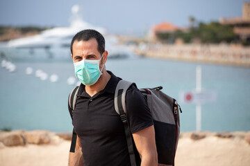 Uomo moro in vacanza con la mascherina chirurgica , maglia nera e zaino in spalla è appena arrivato in una località di mare