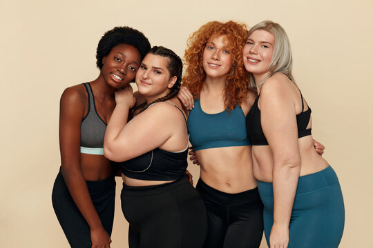 Women. Group Of Diversity Figure And Size Models Portrait. Hugging Multicultural Female In Sportswear Posing Together Against Beige Background. Body Positive As Lifestyle.