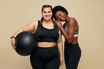 Fototapeta na wymiar Fitness. Slim And Plus Size Models Portrait. Two Different Ethnic Women In Black Sportswear With Pilates Ball Against Beige Background. Sport As Lifestyle.