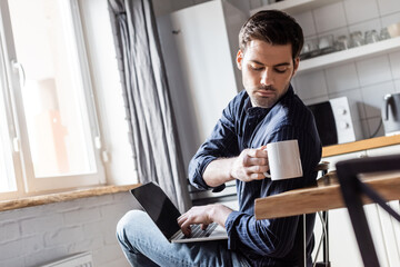 handsome man holding cup of coffee while using laptop on kitchen during quarantine