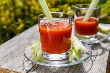 Two glasses of tomato juice with parsley and celery decorations, stand in the open air on wooden boards, the rays of the sun shine, shallow depth of field, selective focus. The concept of natural