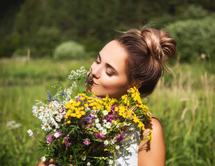 Naklejka premium Summer lifestyle portrait of beautiful young woman smiling and holding bouquet of wild flowers. Standing in the field of grass. Enjoying sun. Happiness and love concept. Сlosed eyes 