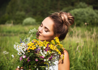 Summer lifestyle portrait  of beautiful young woman smiling and holding bouquet of wild flowers. Standing in the field of grass. Enjoying sun. Happiness and love concept. Сlosed eyes 