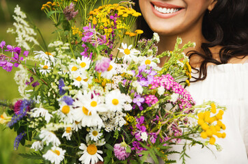 Summer lifestyle portrait of beautiful brunette woman smiling and holding bouquet of wild flowers. Enjoying sun. Happiness and love concept. Beautiful flowers. Romantic mood. Beautiful smile