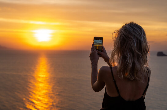 Woman hands holding mobile phone at sunset. Young curly hair woman taking photos with her cell phone in a beautiful amazing sunset over sea. Taking a picture on a smartphone during a vacation