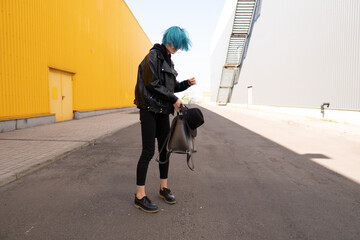 A girl with blue hair in black clothes stands on the street between two buildings and holds a backpack in her hands. - Powered by Adobe