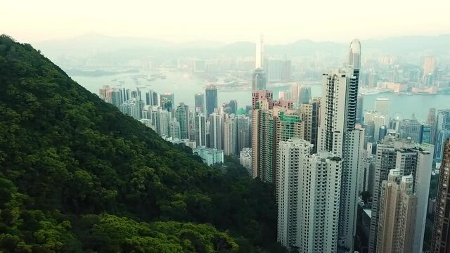 Aerial panning shot of trees on mountain by skyscrapers in city, drone flying over modern buildings against sky at sunset - Hong Kong, China