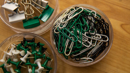 Top view of paper clips, binder and thumbtacks in a transparent box on wooden background. It is device used to hold sheets of paper together made of steel wire bent to a looped shape. Selective focus.