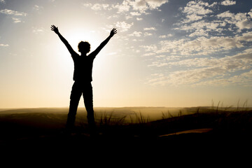 Silhouette of person standing on a dune during sunrise, arms in the air.