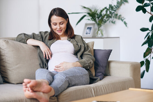 Young barefoot female in casualwear looking at her pregnant belly while relaxing