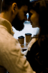 Close up portrait of young romantic couple drinking coffee in paper cups, sitting in a cozy cafe, touching foreheads and enjoying moments together. Focus on cups of coffee on the table