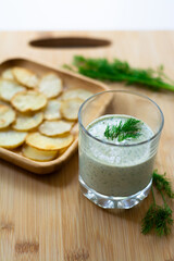 Dill and sour cream dip for homemade potato chips made vegan: healthy snack with dill, tahini, water, vinegar and lemon juice in a glass. Thinly sliced and baked potatoes in the back