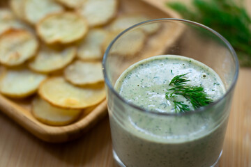 Dill and sour cream dip for homemade potato chips made vegan: healthy snack with dill, tahini, water, vinegar and lemon juice in a glass. Thinly sliced and baked potatoes in the back