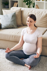Young positive pregnant woman in activewear sitting in pose of lotus by couch