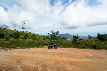 Blue scooter on the dirt road on a hill in tropical jungle with a scenic view on a mountains in a sunny day. Adventure and traveling concept.