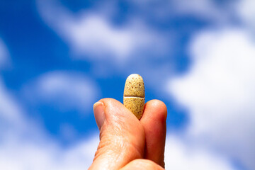 A vitamin tablet or pill held in a hand in front of a cloudy sky