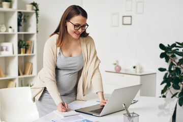 Pregnant businesswoman in smart casualwear bending over desk by laptop