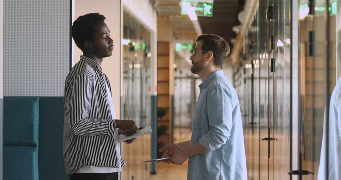 Diverse male partners talking handshaking standing in office hallway, satisfied african american business man shaking hand of caucasian colleague thanking for help advice meeting in company corridor