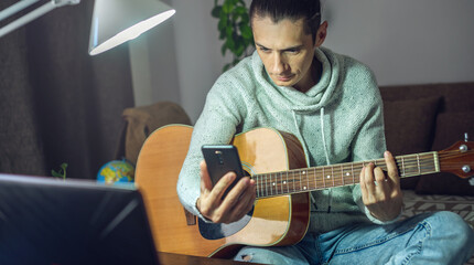Young male musician is learning to play acoustic guitar in an online lesson using a laptop at night in a room at home