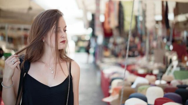 Pretty long red hair girl walking on local flea market and watching different handmade products. Assortment of summer hats displayed on market counter. Trading on local market