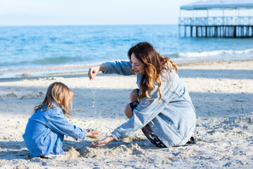 Fototapeta na wymiar young woman in jeans jacket playing with little girl in sand on the beach