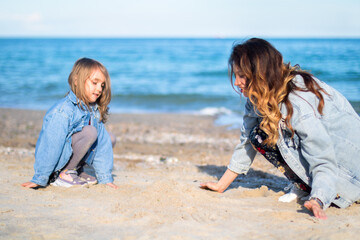 Fototapeta na wymiar young woman in jeans jacket playing with little girl in sand on the beach